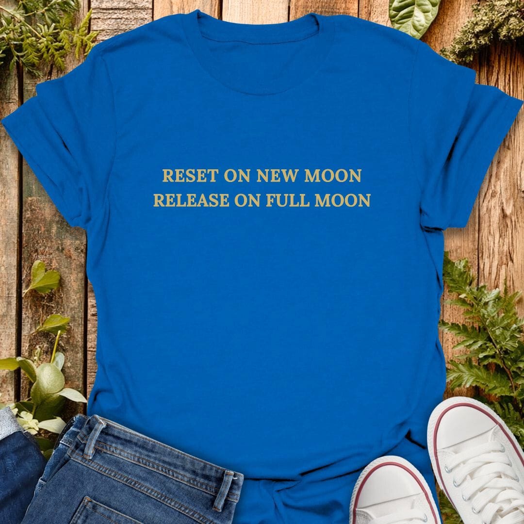 Blue t-shirt with text 'Reset on new moon, release on full moon' on a wooden surface with plants, jeans and sneakers.