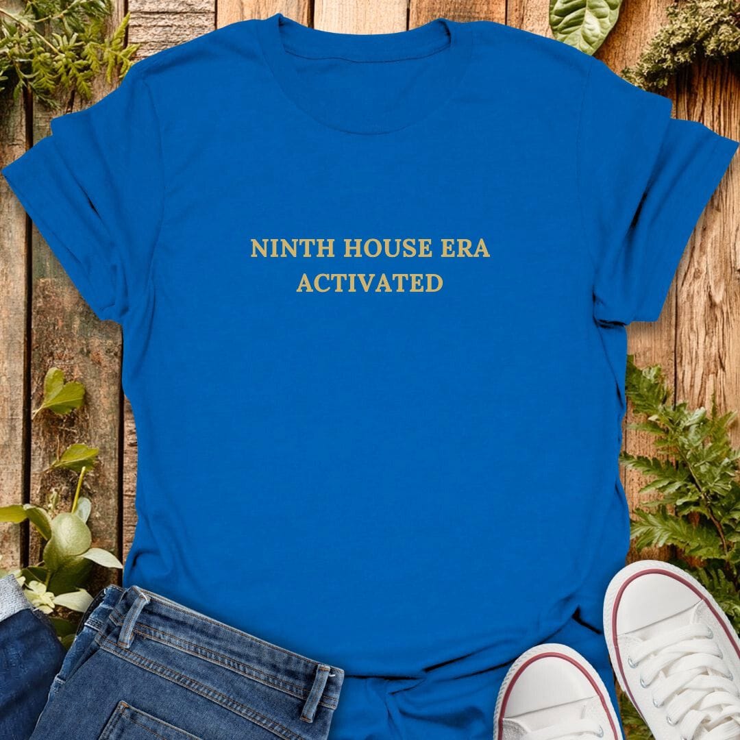 Blue t-shirt with 'Ninth house era activated' text on a wooden surface with plants, jeans and sneakers.