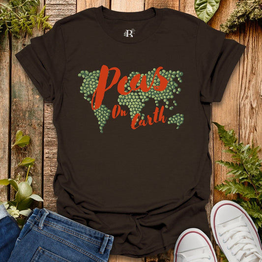 Black t-shirt with 'Peas on Earth' design on a wooden surface with plants.