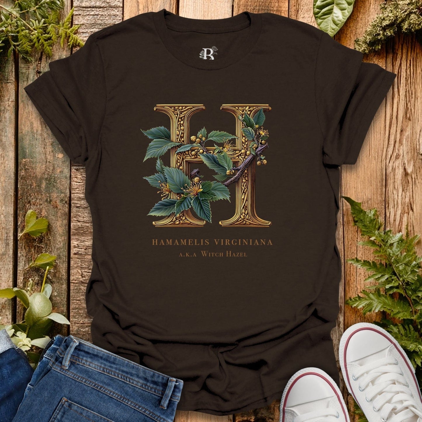 Black t-shirt with gold letter 'H' and plant design on a wooden surface with plants around.
