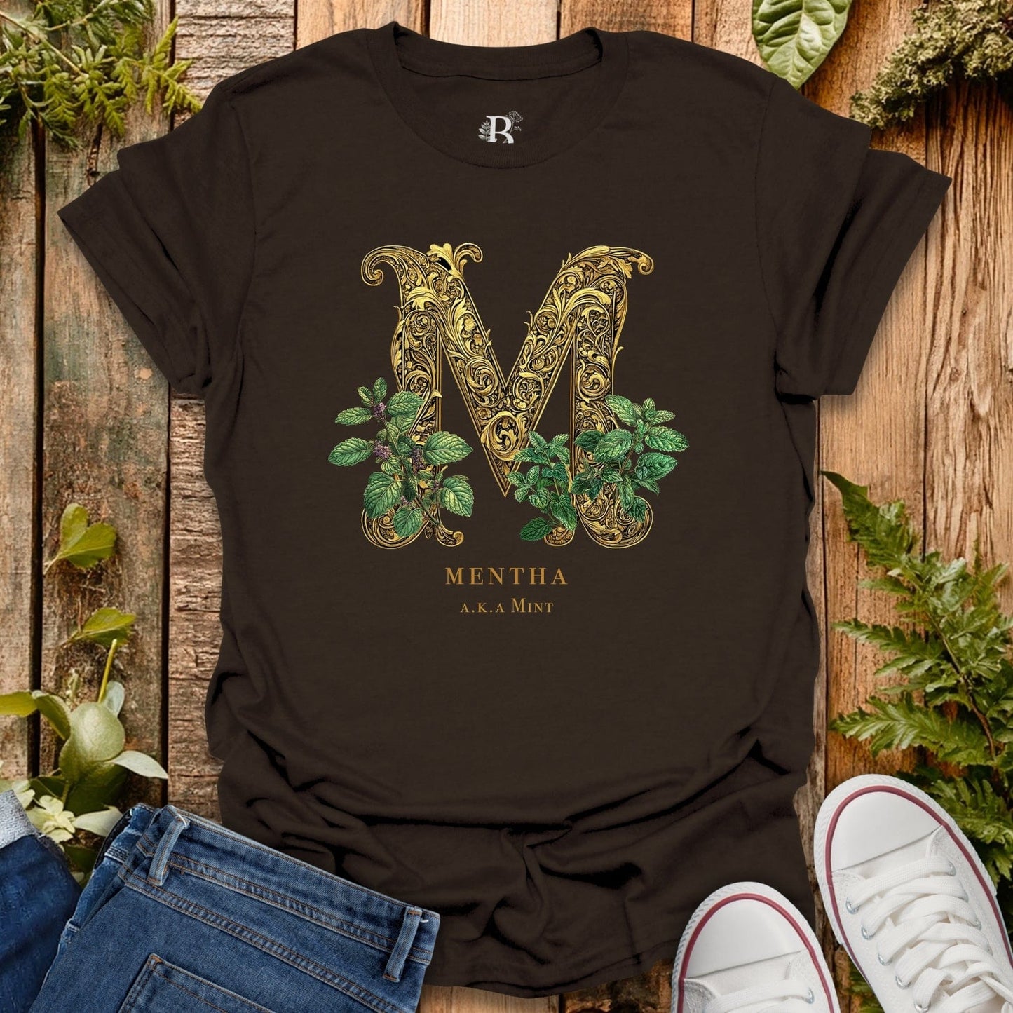 Black t-shirt with decorative letter 'M' and plant design on a wooden surface with plants around.