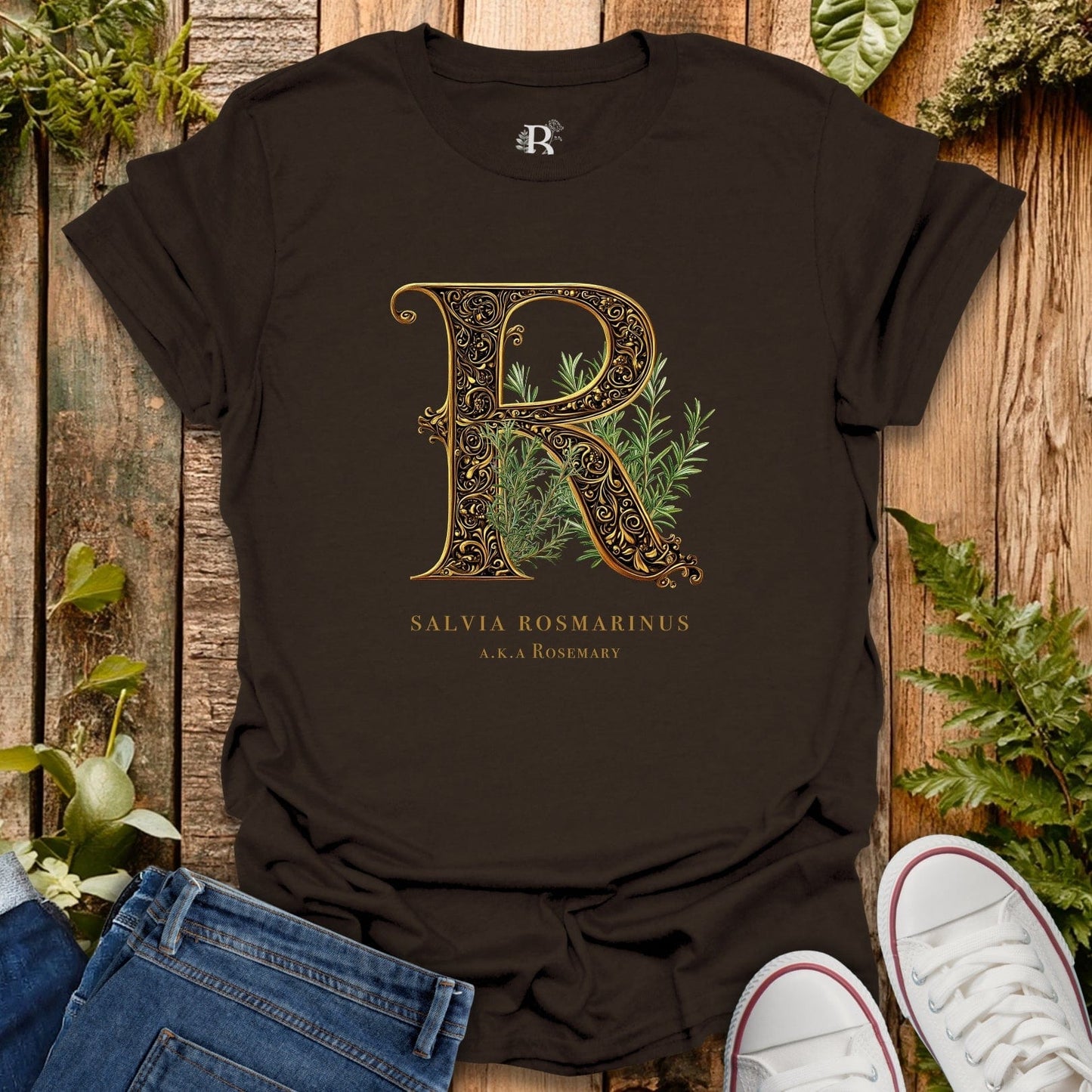 Black t-shirt with decorative letter 'R' and plant design on a wooden surface with plants around.