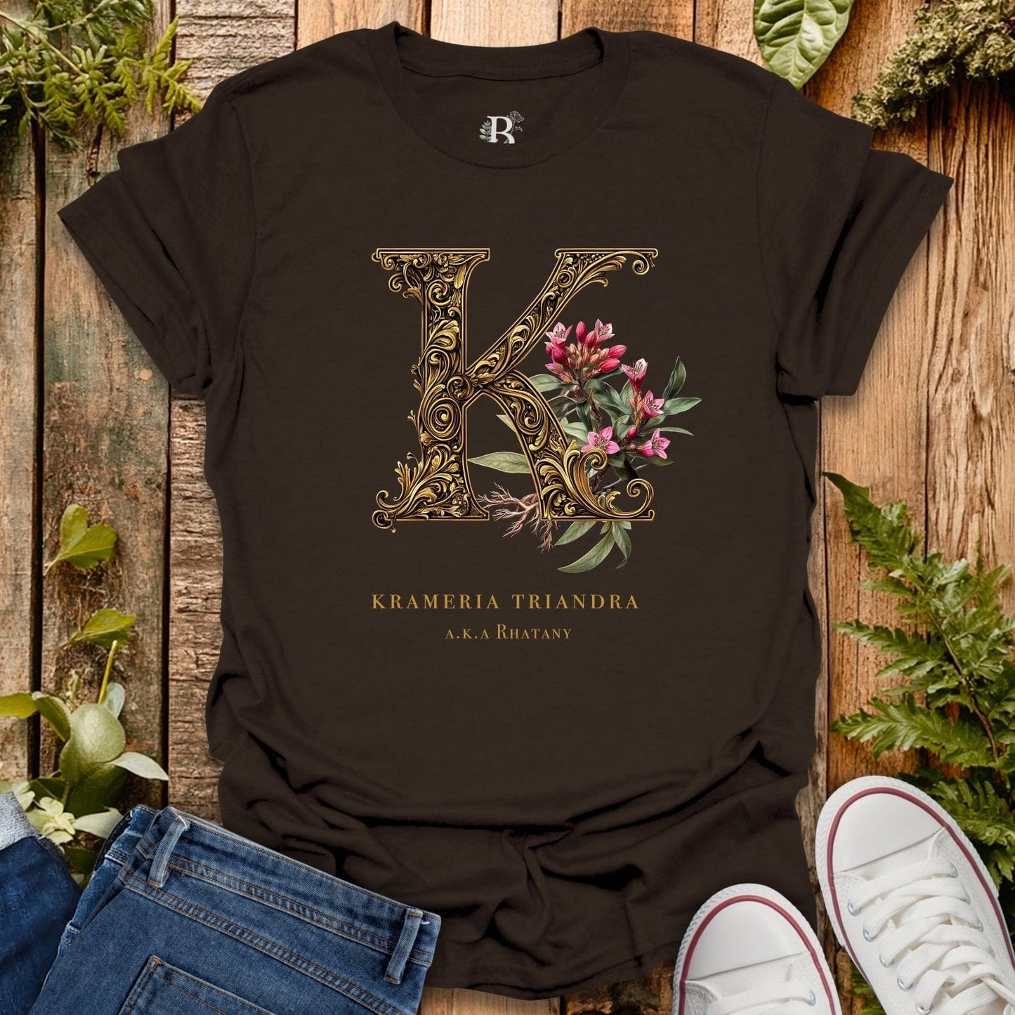 Black t-shirt with decorative letter 'K' and floral design on a wooden surface with plants.