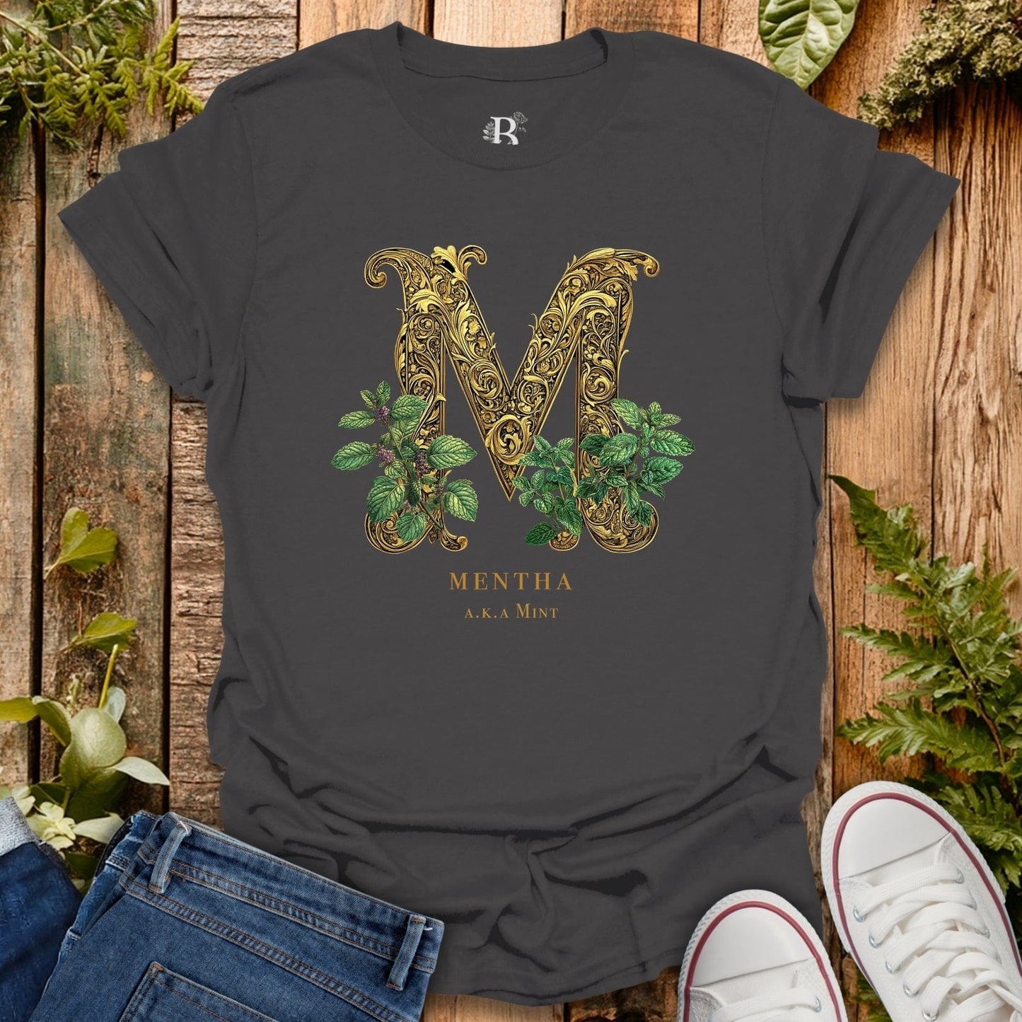 Black t-shirt with decorative letter 'M' and plant design on a wooden surface with plants around.