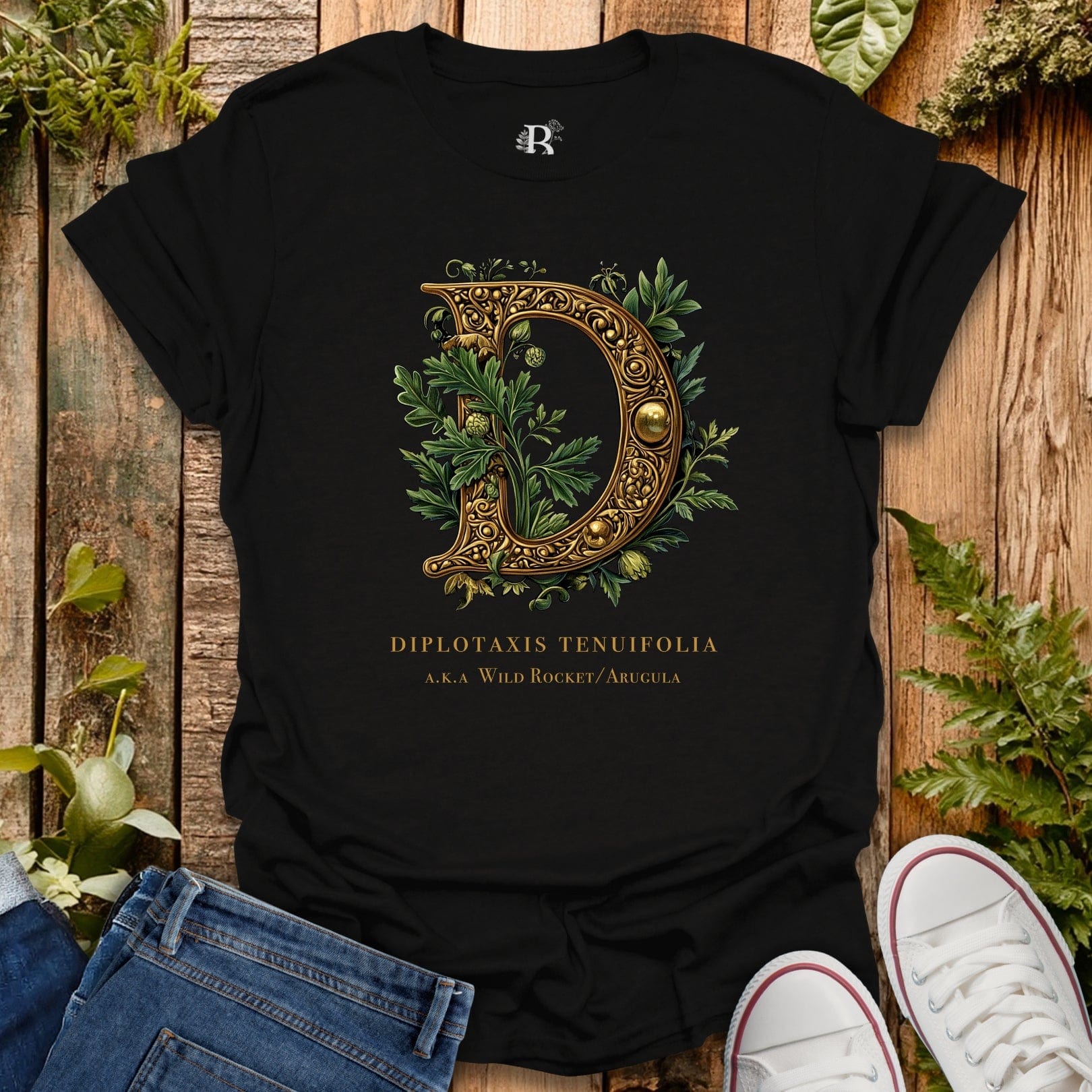 Black t-shirt with decorative letter 'D' and plant design on a wooden surface with plants.