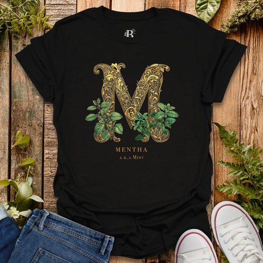 Black t-shirt with decorative letter 'M' and plant design on a wooden surface with plants around.