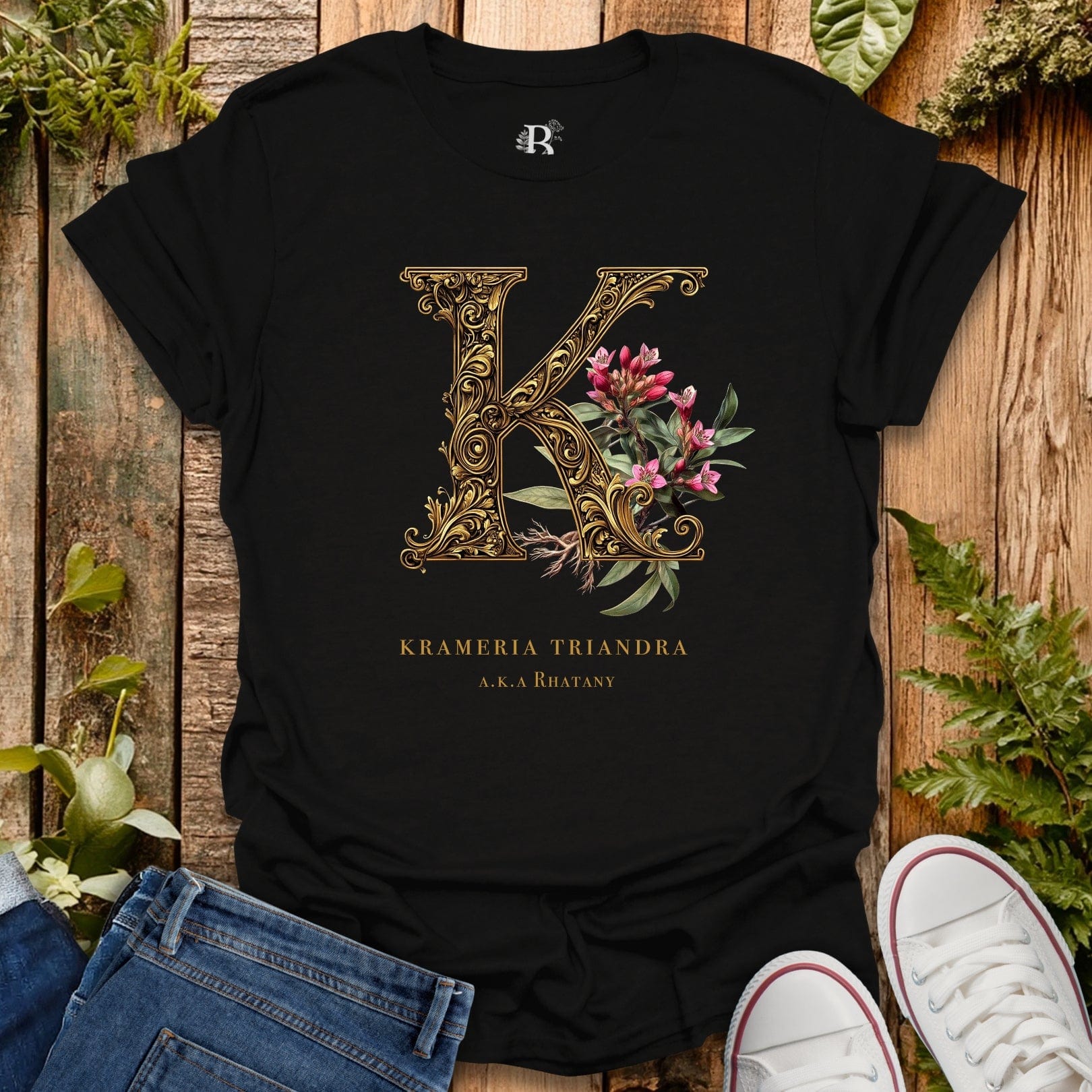 Black t-shirt with ornate letter 'K' design on a wooden surface with plants.