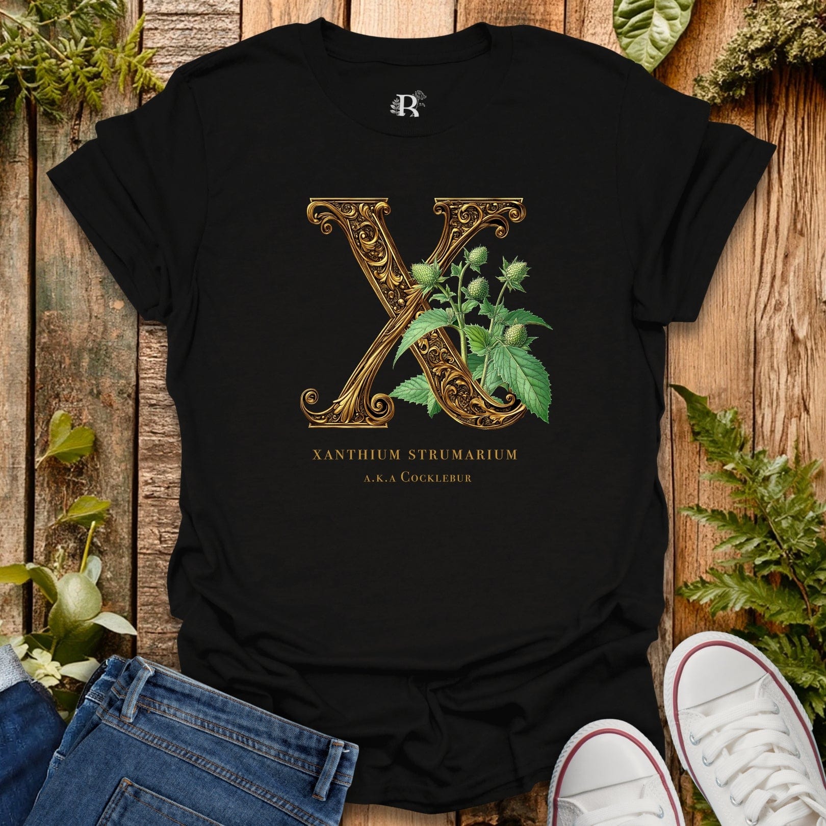 Black t-shirt with gold letter 'X' and plant design on a wooden surface with plants.