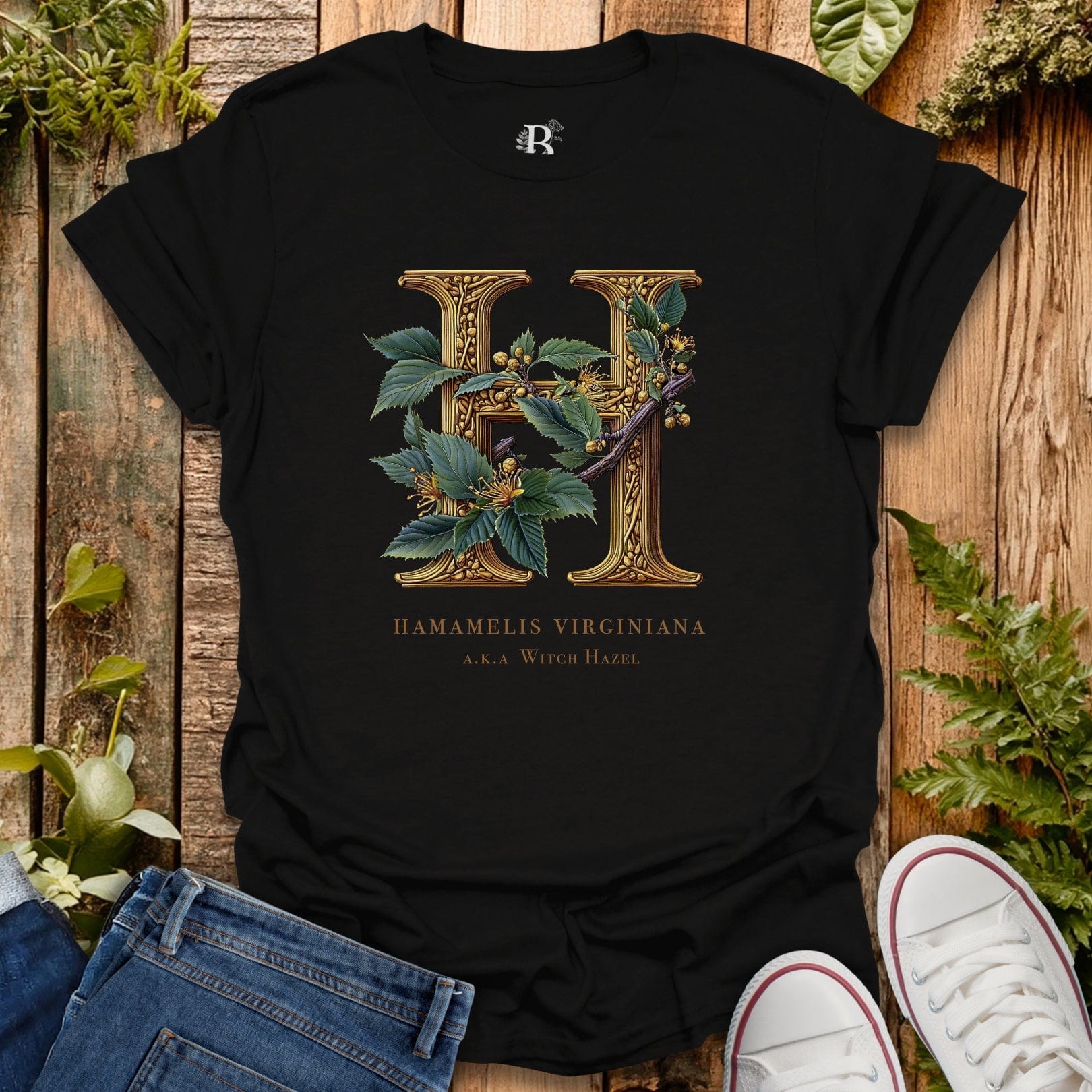Black t-shirt with gold letter 'H' and plant design on a wooden surface with plants.