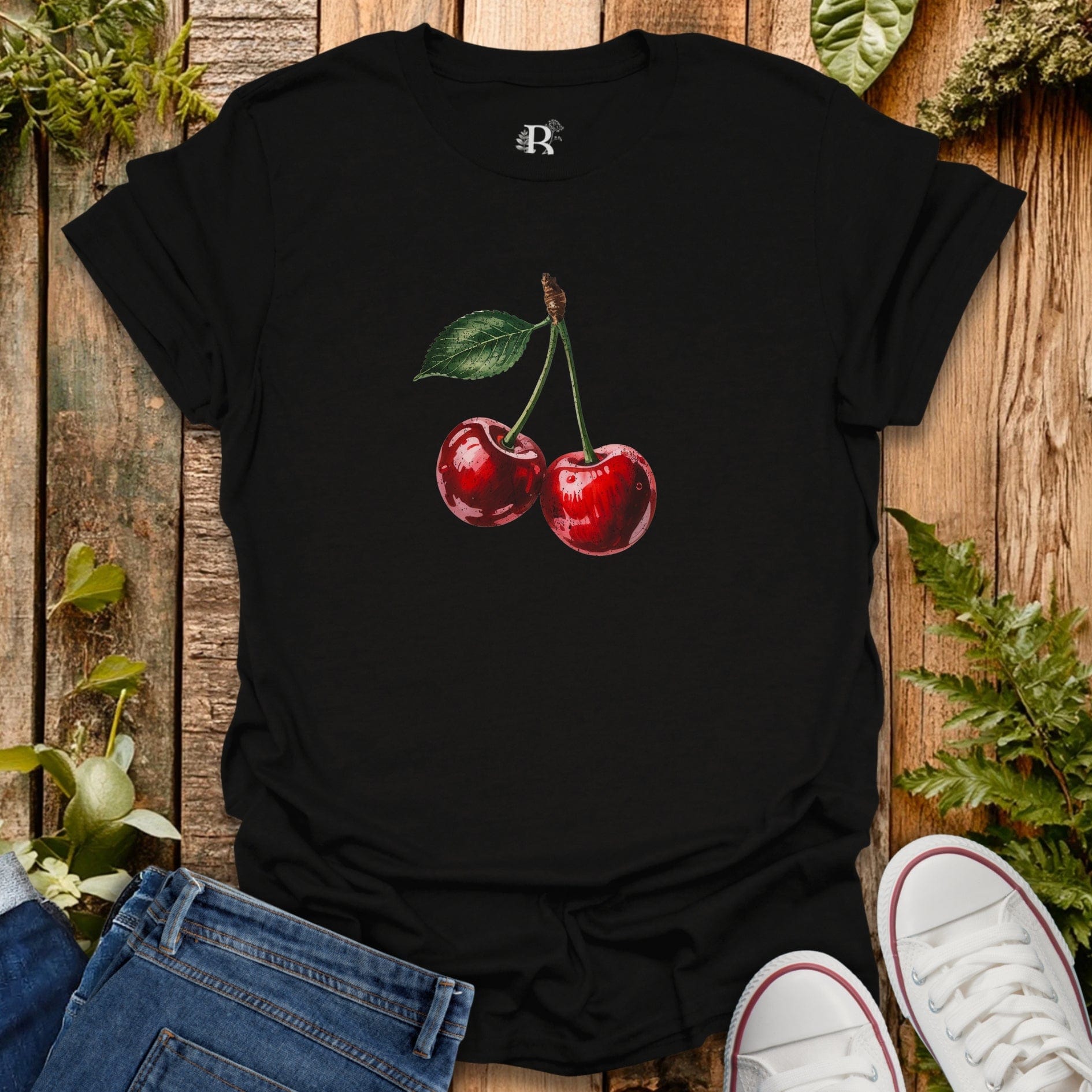 Black t-shirt with cherry design on a wooden surface with jeans and sneakers.