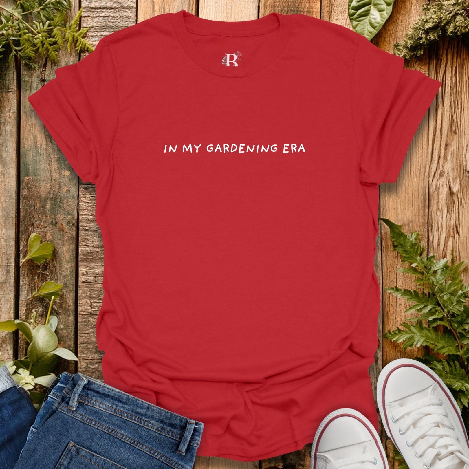 Red t-shirt with 'IN MY GARDENING ERA' text on a wooden surface with plants.