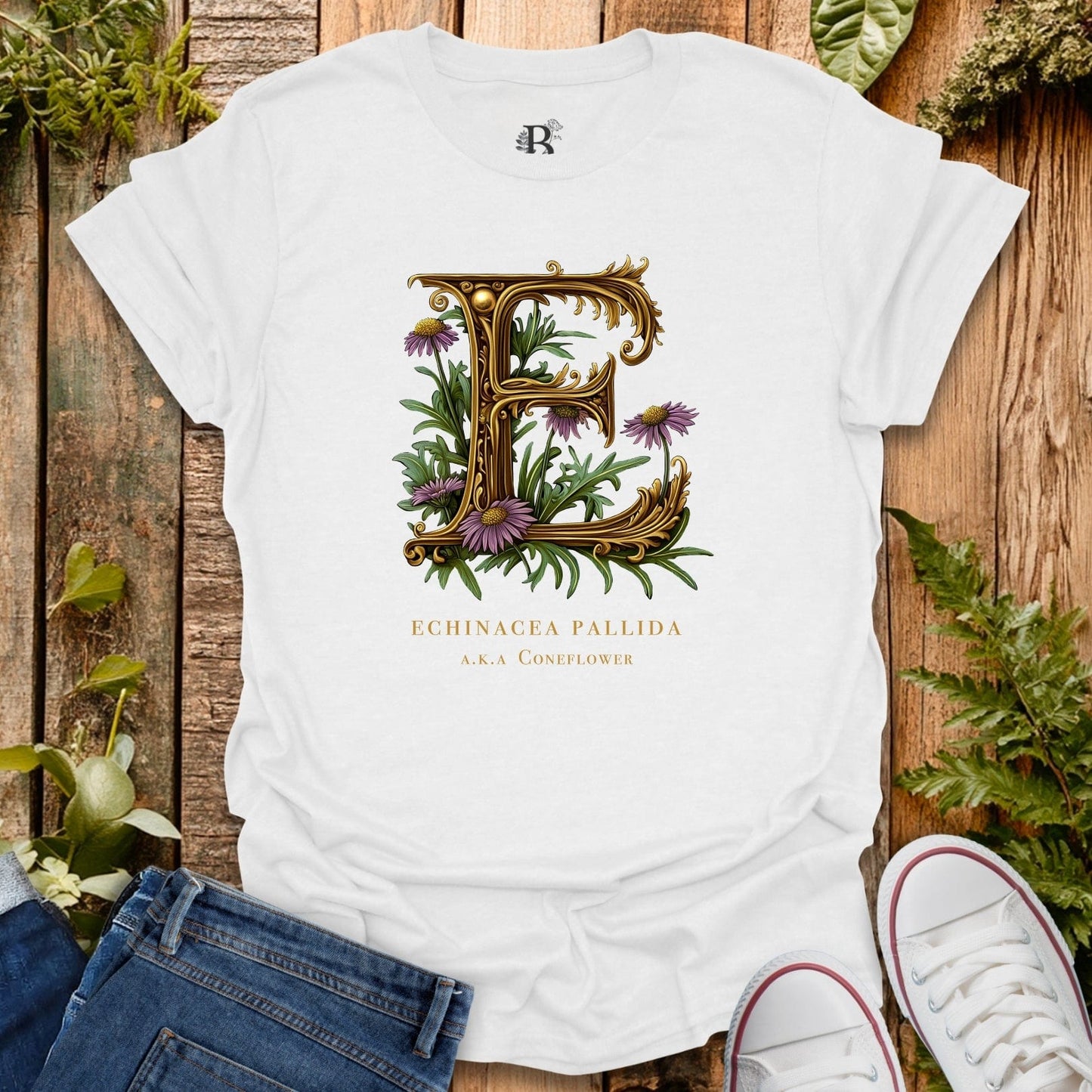 White t-shirt with decorative letter 'E' design featuring flowers on a wooden surface with plants.