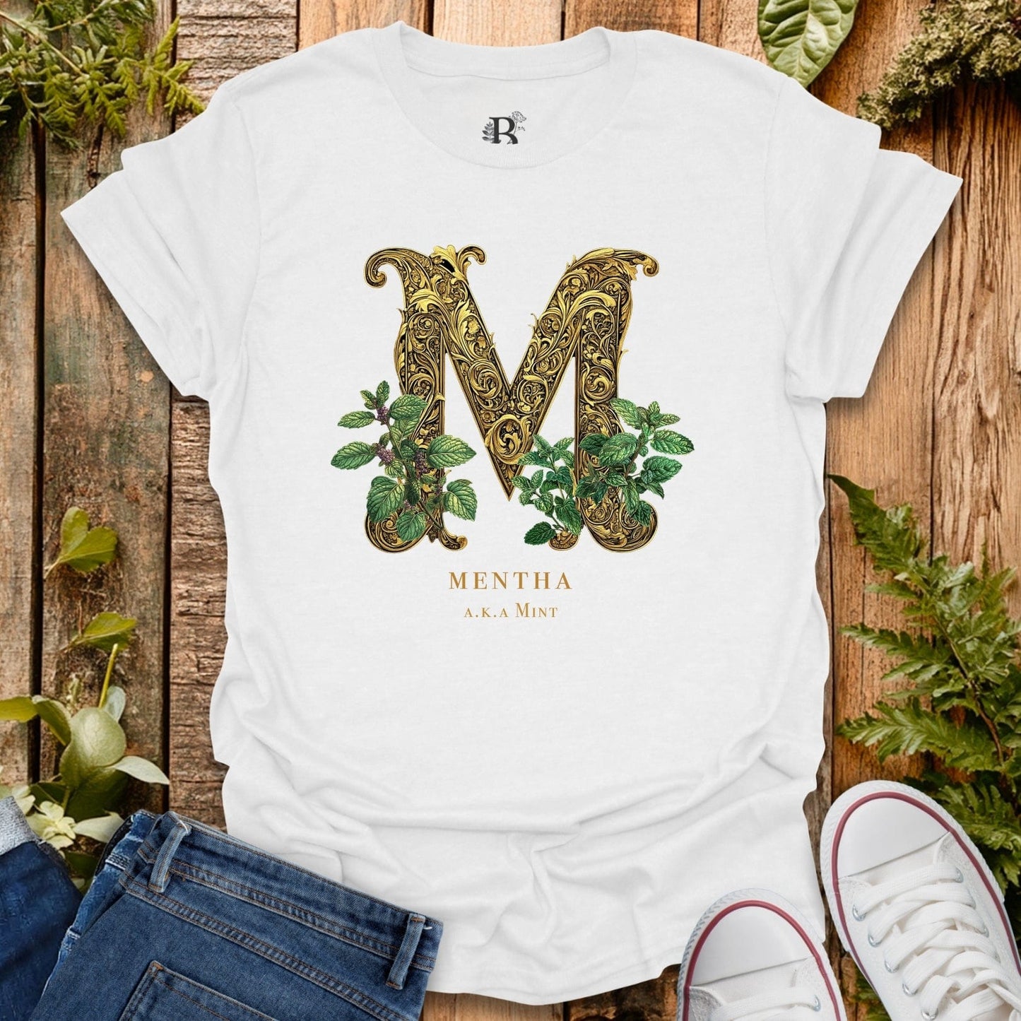 White t-shirt with decorative letter 'M' and plant design on a wooden surface with plants around.
