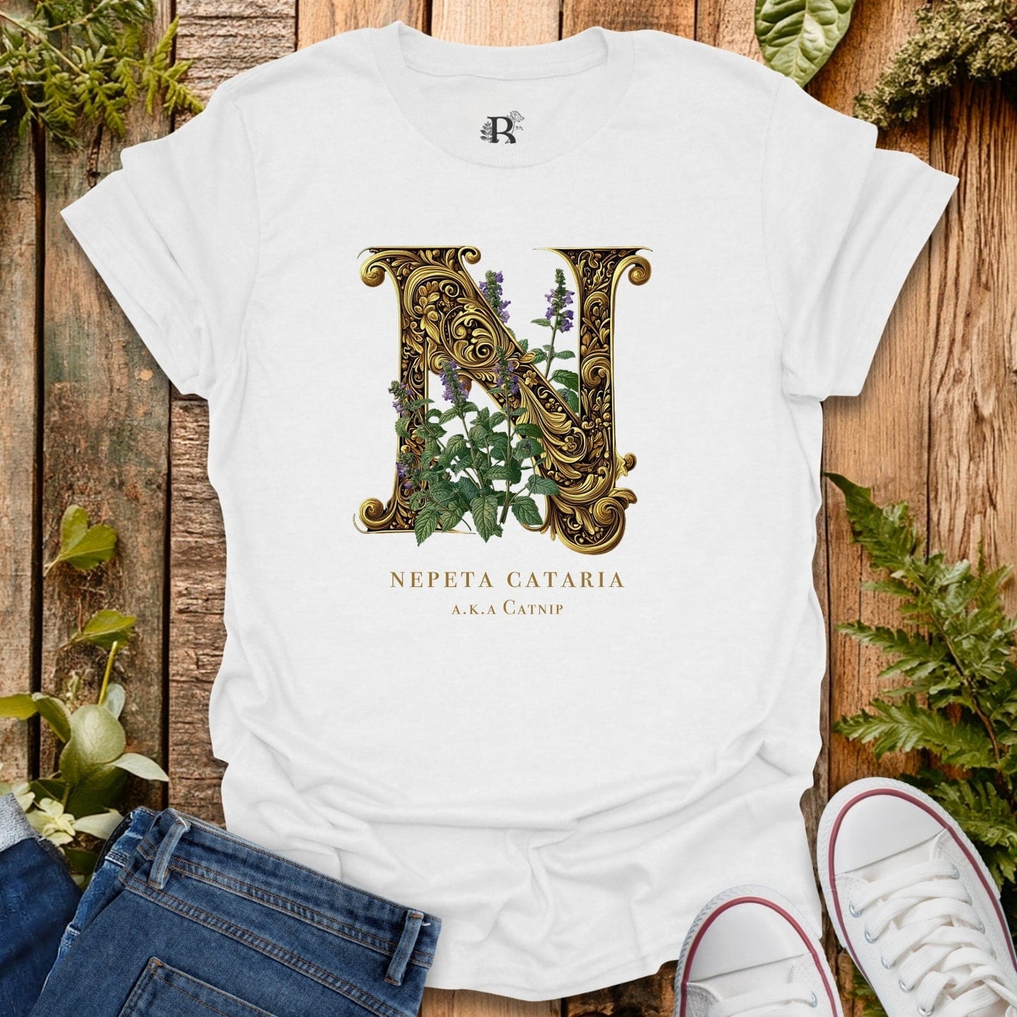 White t-shirt with decorative letter 'N' and plant design on a wooden surface with plants around.