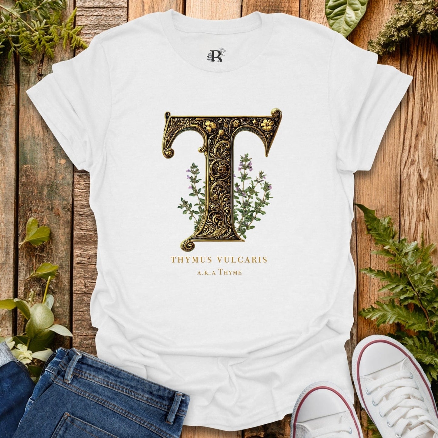 White t-shirt with decorative letter 'T' and plant design on a wooden surface with plants.