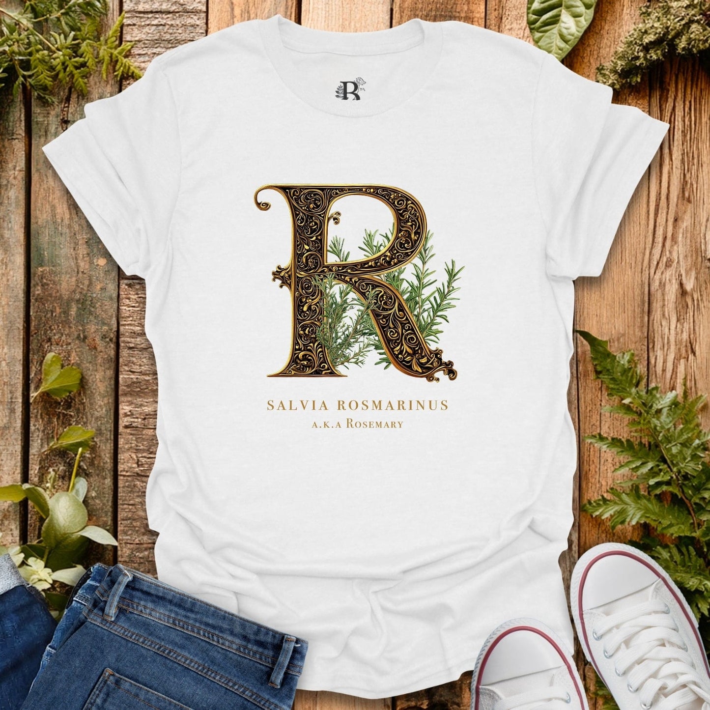 White t-shirt with decorative letter 'R' and plant design on a wooden surface with plants around.