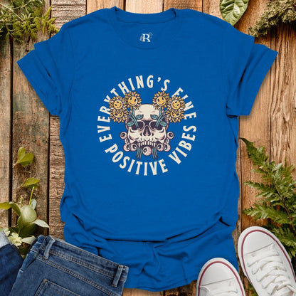 Blue t-shirt with skull and flowers design on a wooden surface with jeans and sneakers.