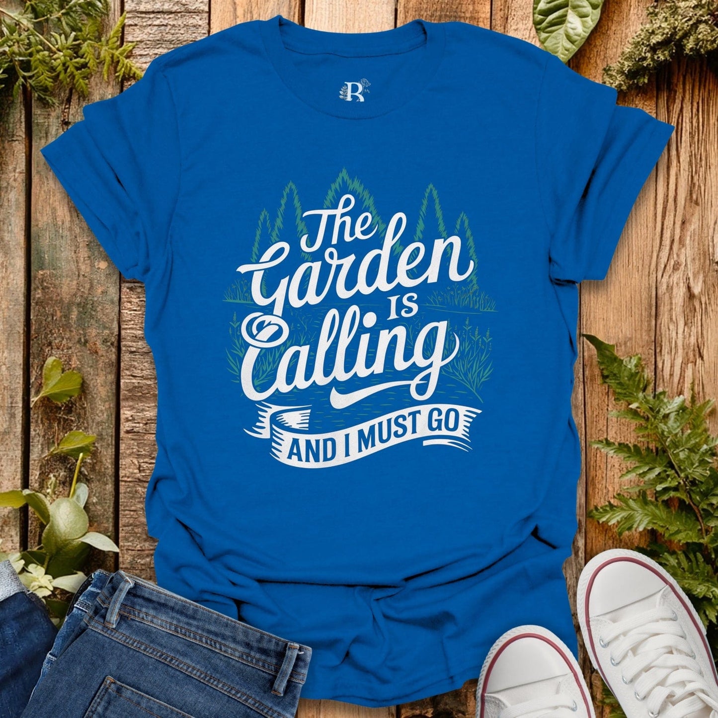 Blue t-shirt with 'The Garden is Calling and I Must Go' text on a wooden background with plants.