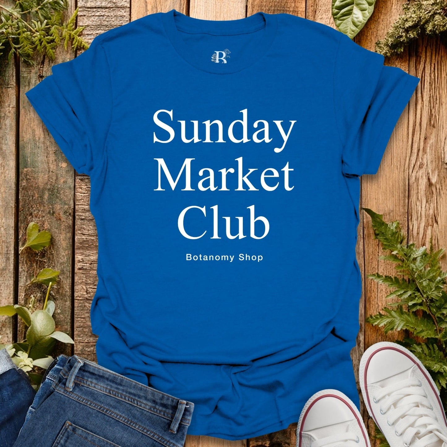 Blue t-shirt with 'Sunday Market Club' text on a wooden surface with plants.