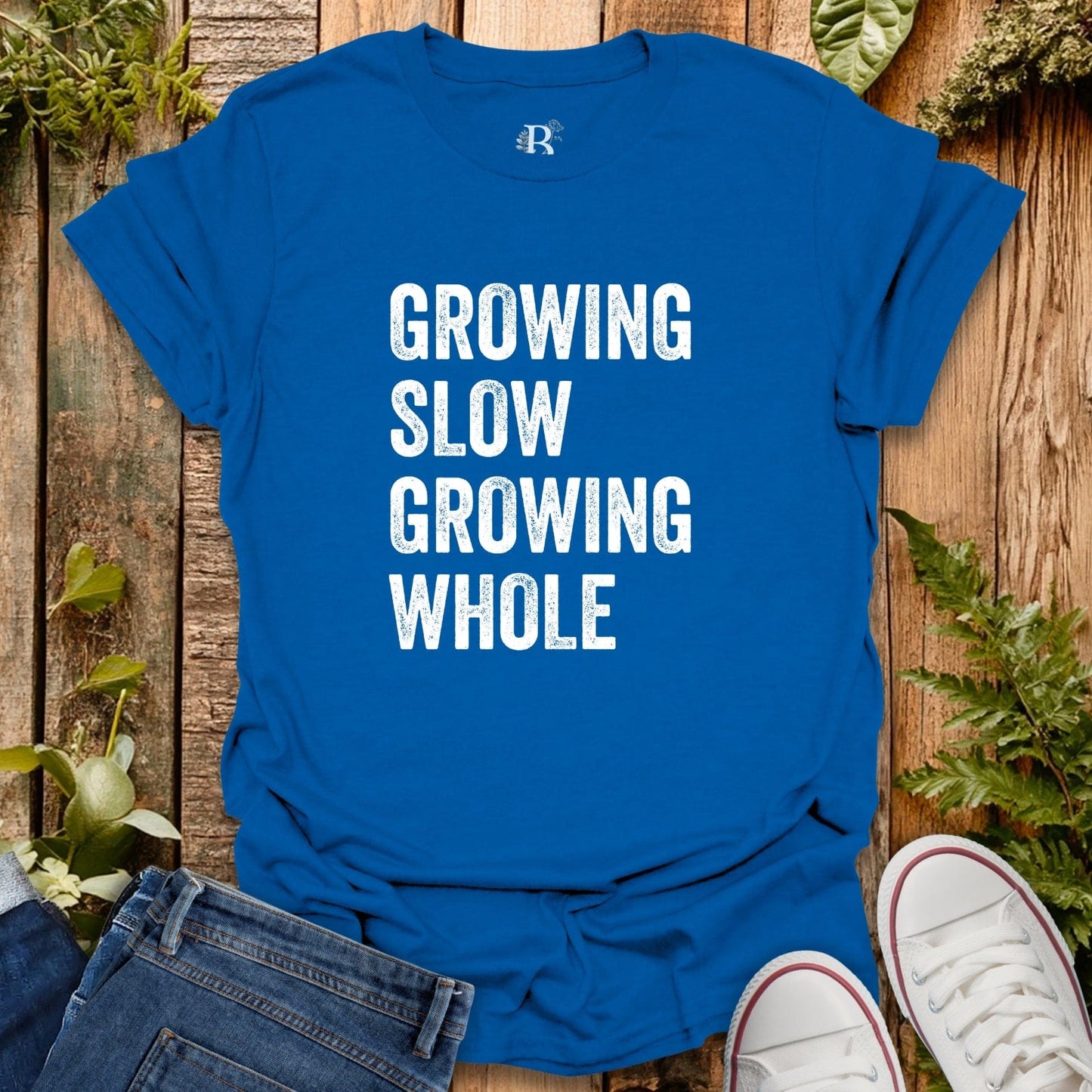 Blue t-shirt with 'GROWING SLOW GROWING WHOLE' text on a wooden surface with plants.