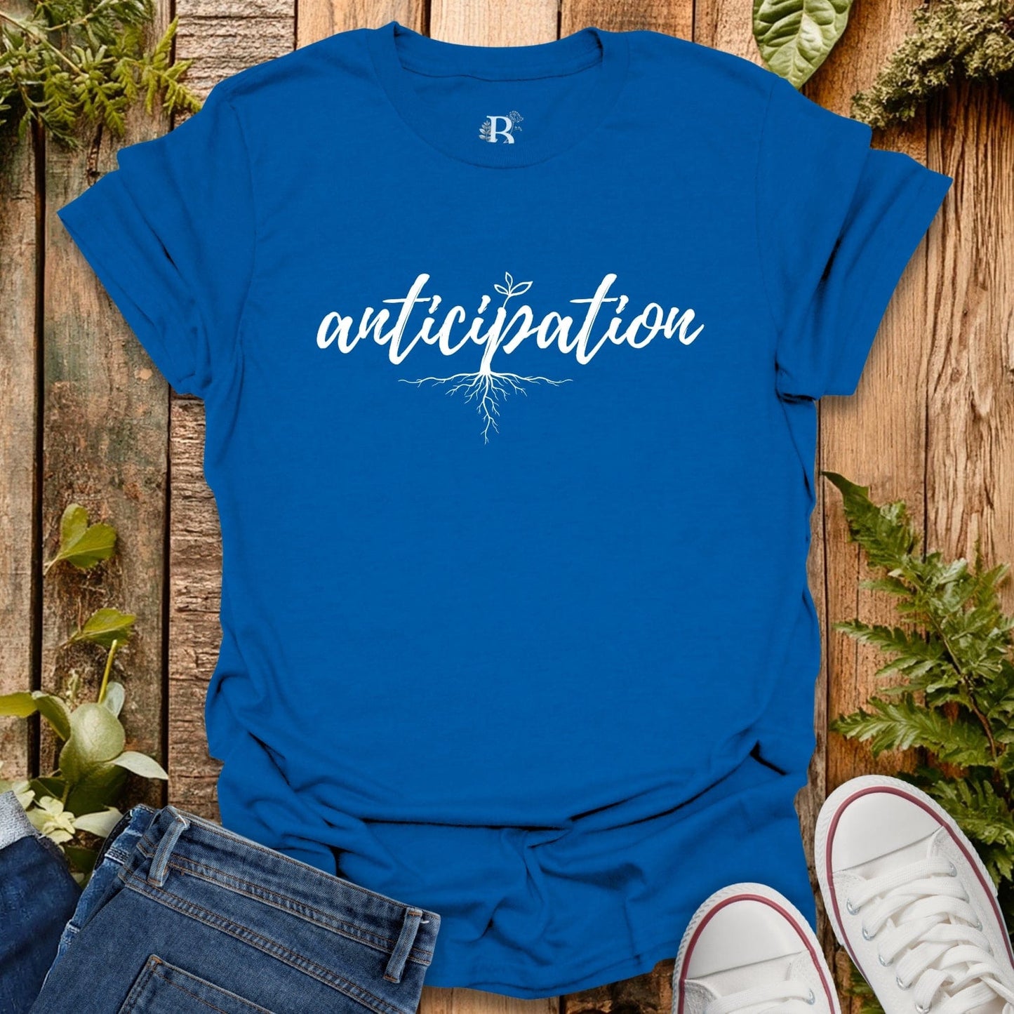 Blue t-shirt with 'anticipation' text on a wooden surface with plants.