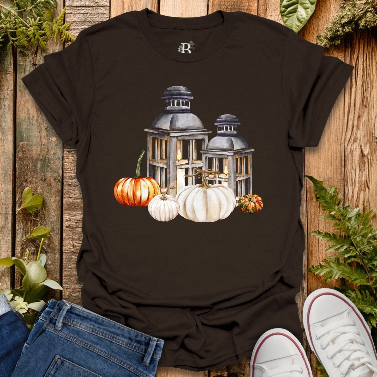 Brown t-shirt with lantern and pumpkin design on a wooden surface with jeans and sneakers.
