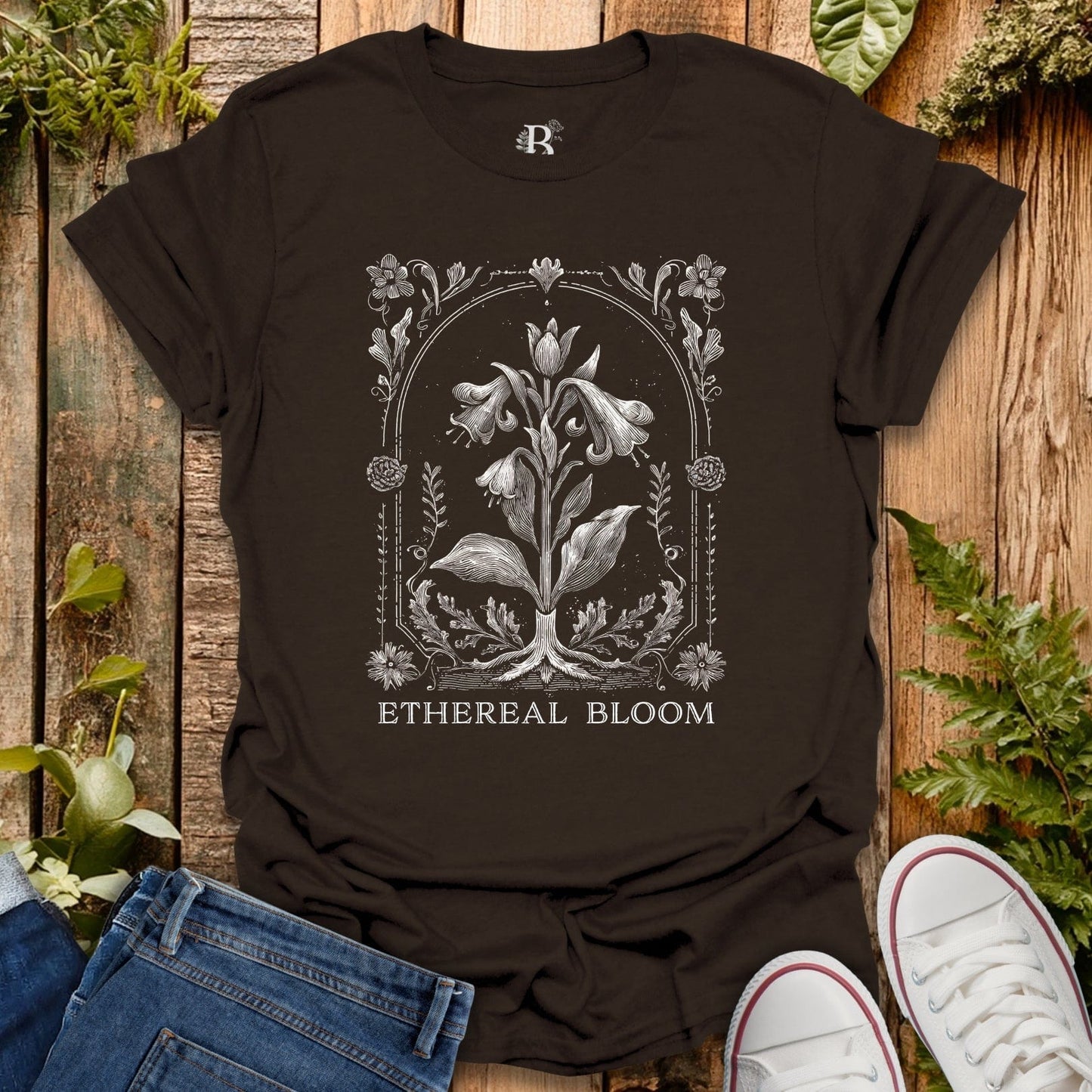 Brown t-shirt with floral design and 'Ethereal Bloom' text on a wooden surface with plants.