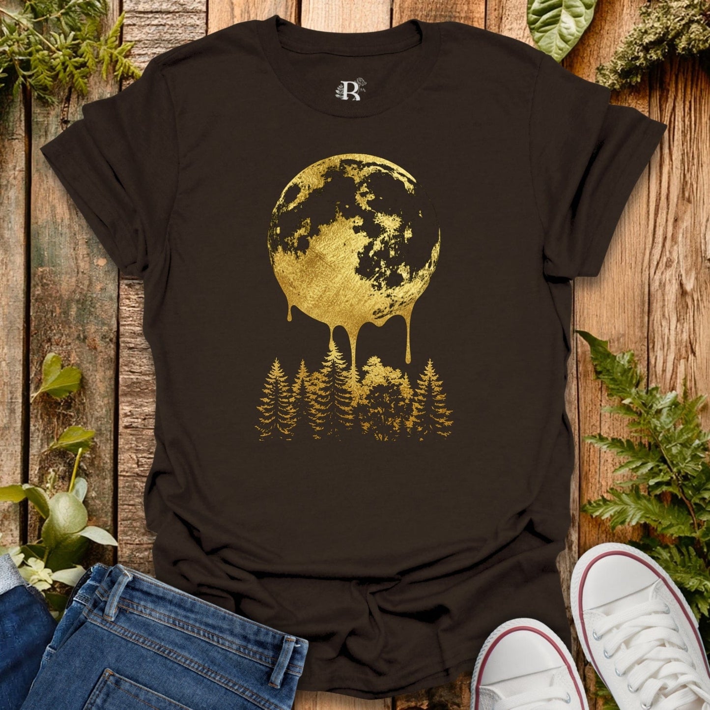 Brown t-shirt with gold moon and forest design on a wooden surface with plants.