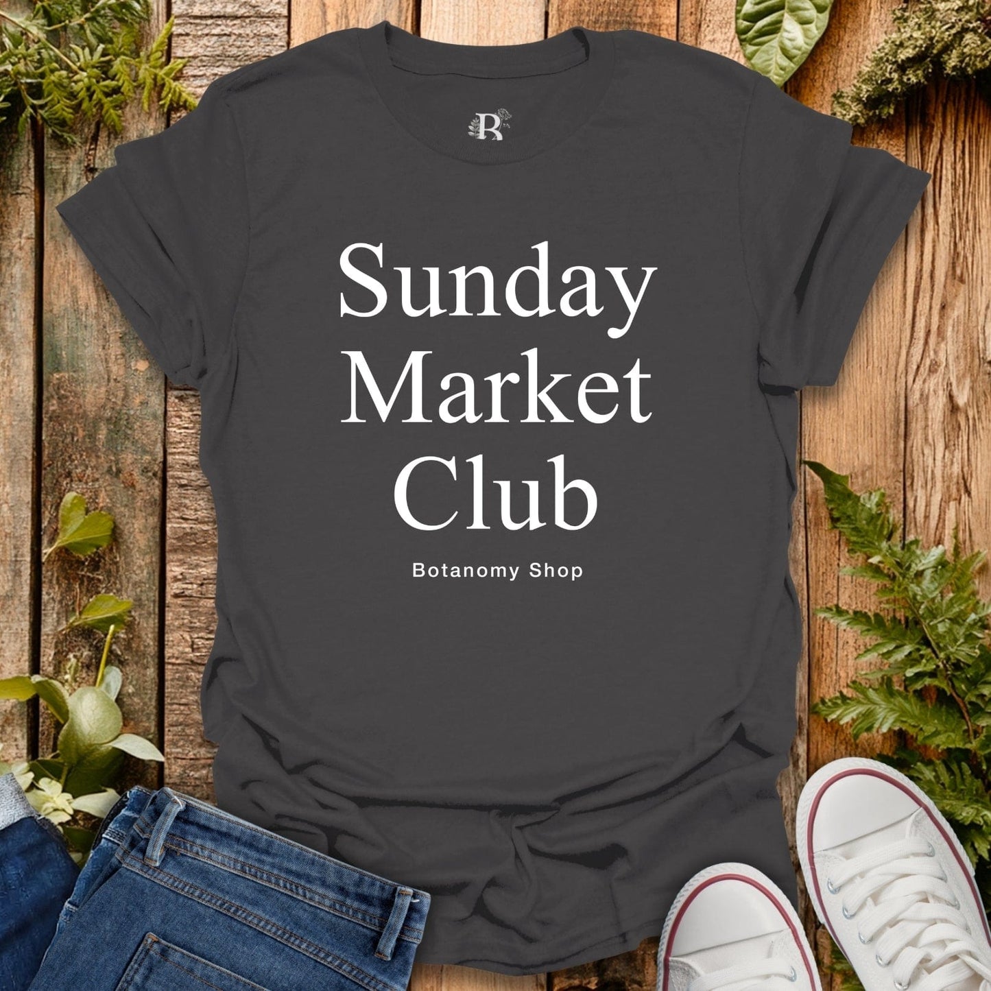 Black t-shirt with 'Sunday Market Club' text on a wooden surface with plants.