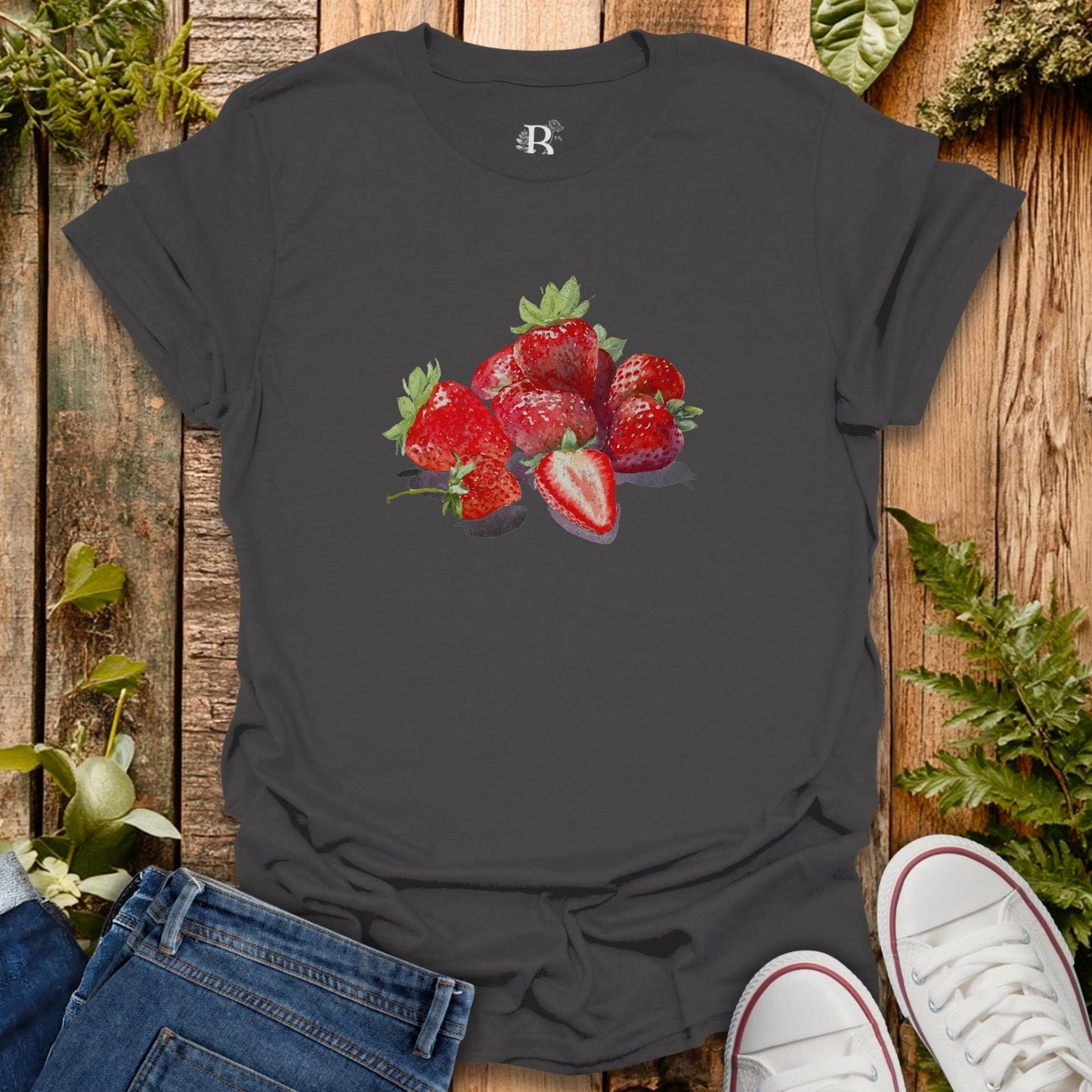 Dark gray t-shirt with strawberry design on a wooden surface with jeans and sneakers.