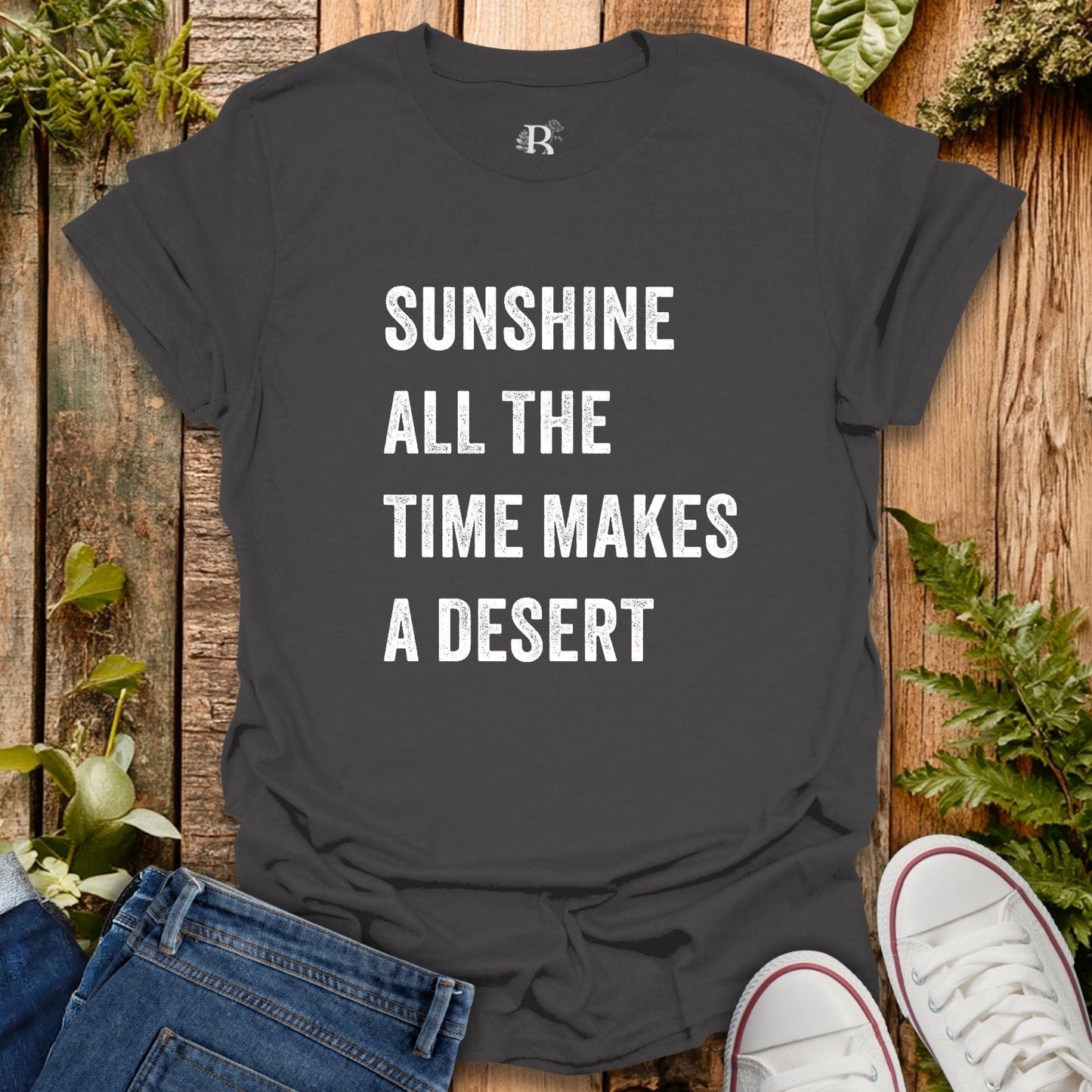 Black t-shirt with 'Sunshine all the time makes a desert' text on a wooden surface with plants.