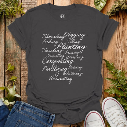 Dark gray t-shirt with gardening-related text on a wooden surface with plants.