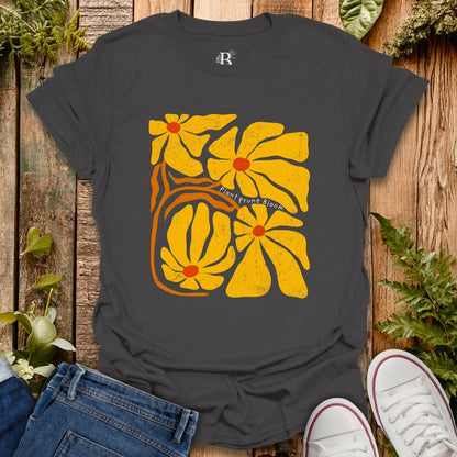 Black t-shirt with yellow floral design on a wooden surface with plants.