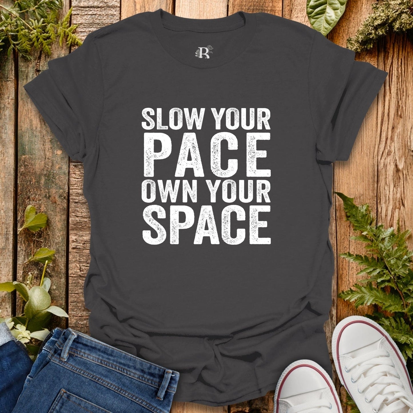 Black t-shirt with 'Slow your pace, own your space' text on a wooden background with jeans and sneakers.