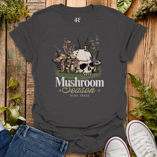 Dark gray t-shirt with mushroom graphic and text on a wooden surface with plants.
