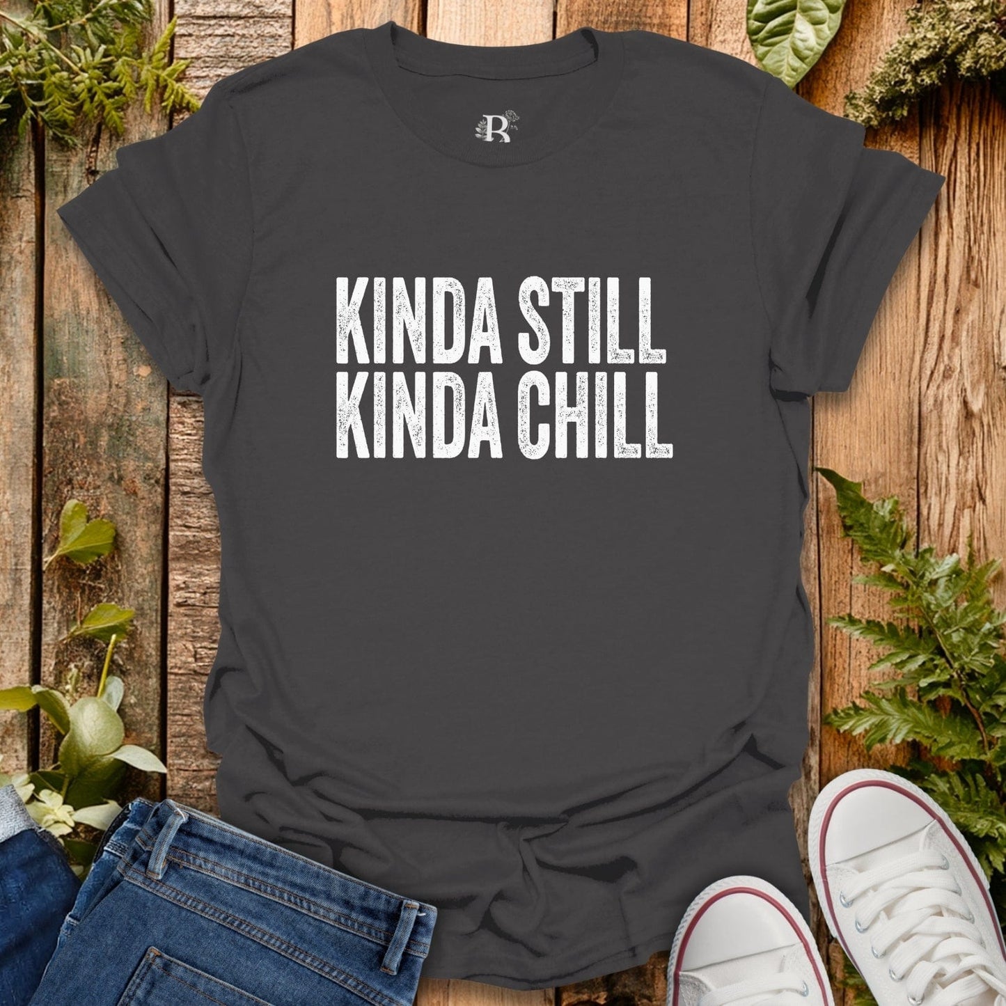 Black t-shirt with 'KINDA STILL KINDA CHILL' text on a wooden surface with jeans and sneakers.