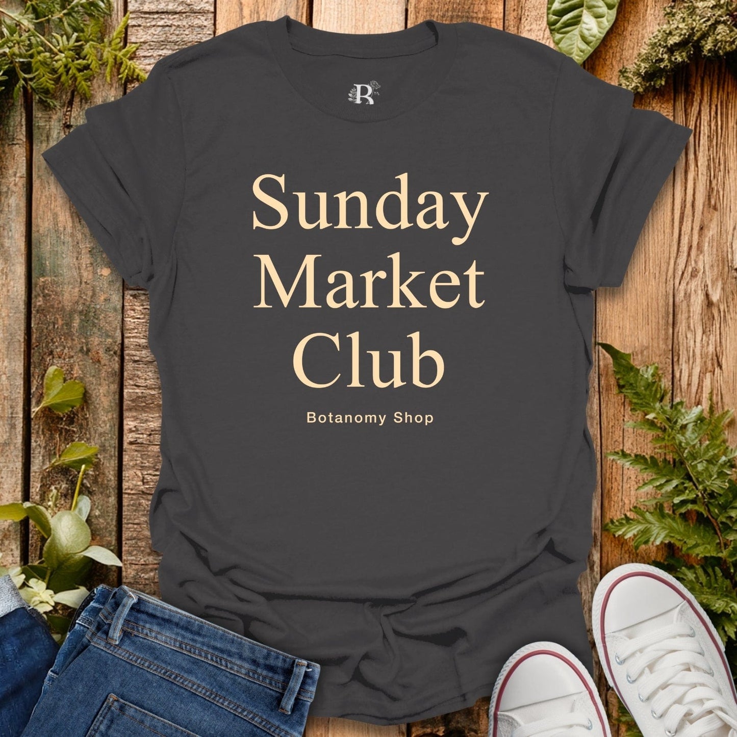 Black t-shirt with 'Sunday Market Club' text on a wooden surface with plants.