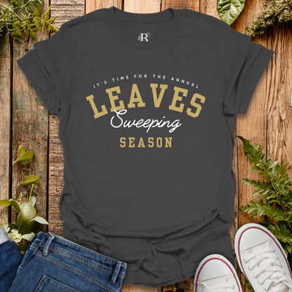 Dark gray t-shirt with 'Leaves Sweeping Season' text on a wooden background with shoes and leaves.