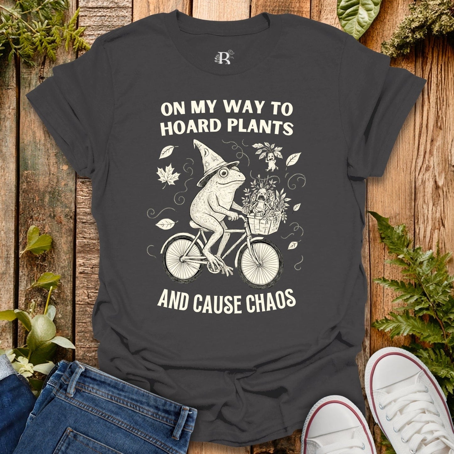 Gray t-shirt with a graphic of a frog riding a bike and text on a wooden background with plants.
