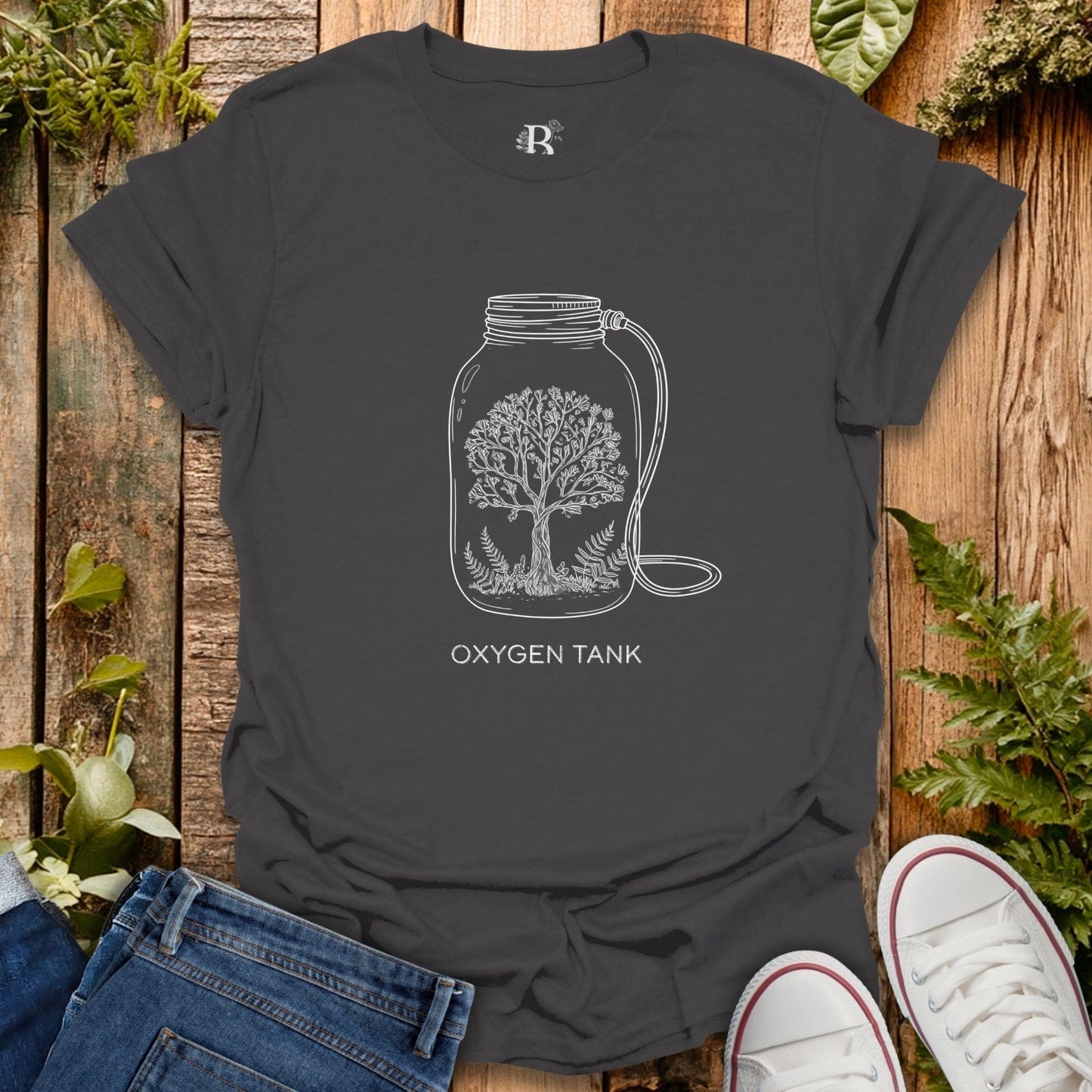 Dark gray t-shirt with a tree design inside a jar labeled 'Oxygen Tank' on a wooden surface with plants.