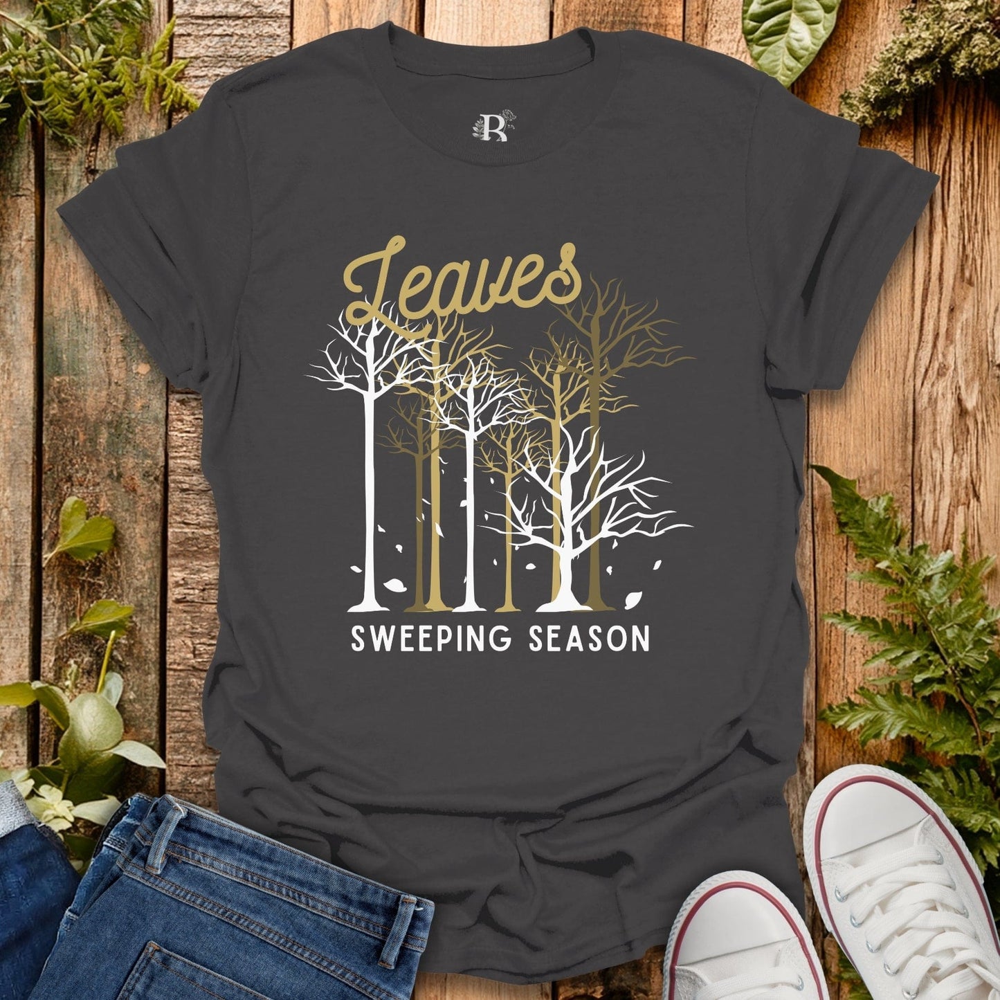 Black t-shirt with 'Leaves Sweeping Season' design on a wooden surface with plants.