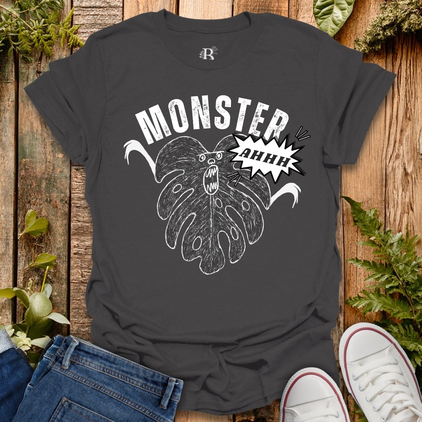 Gray t-shirt with a graphic design of a monster and the word 'MONSTER' on a wooden surface with plants.