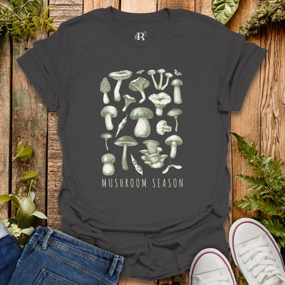 Black t-shirt with green mushroom illustrations and 'Mushroom Season' text on a wooden surface with plants.