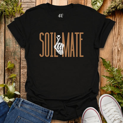 Black t-shirt with 'SOIL MATE' text and graphic on a wooden surface with plants.