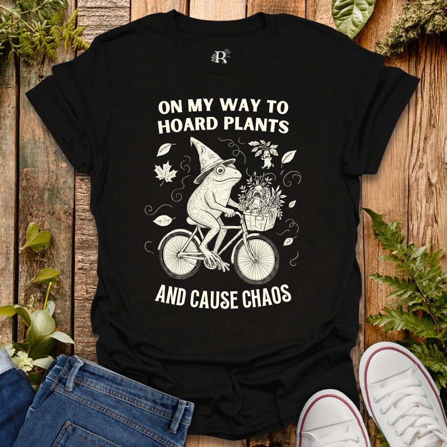 Black t-shirt with a graphic of a frog riding a bike and text on a wooden background with plants.