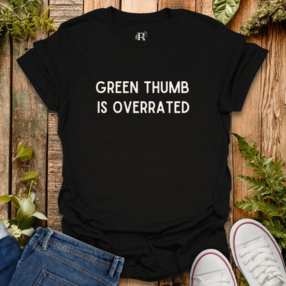 Black t-shirt with 'Green Thumb is Overrated' text on a wooden surface with plants.