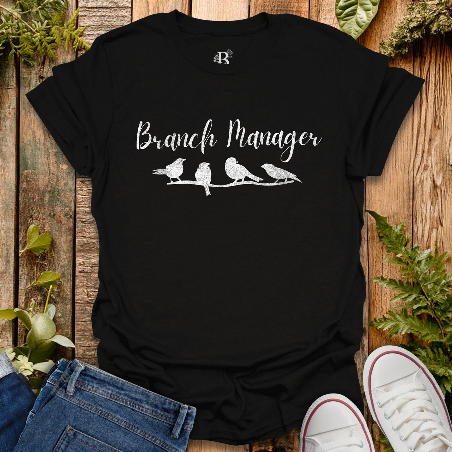 Black t-shirt with 'Branch Manager' text and bird illustrations on a wooden surface with plants.