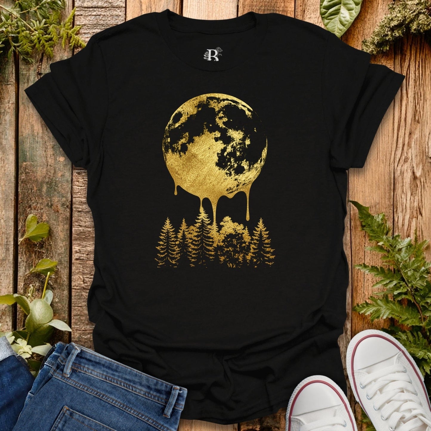 Black t-shirt with gold moon and forest design on a wooden surface with plants.
