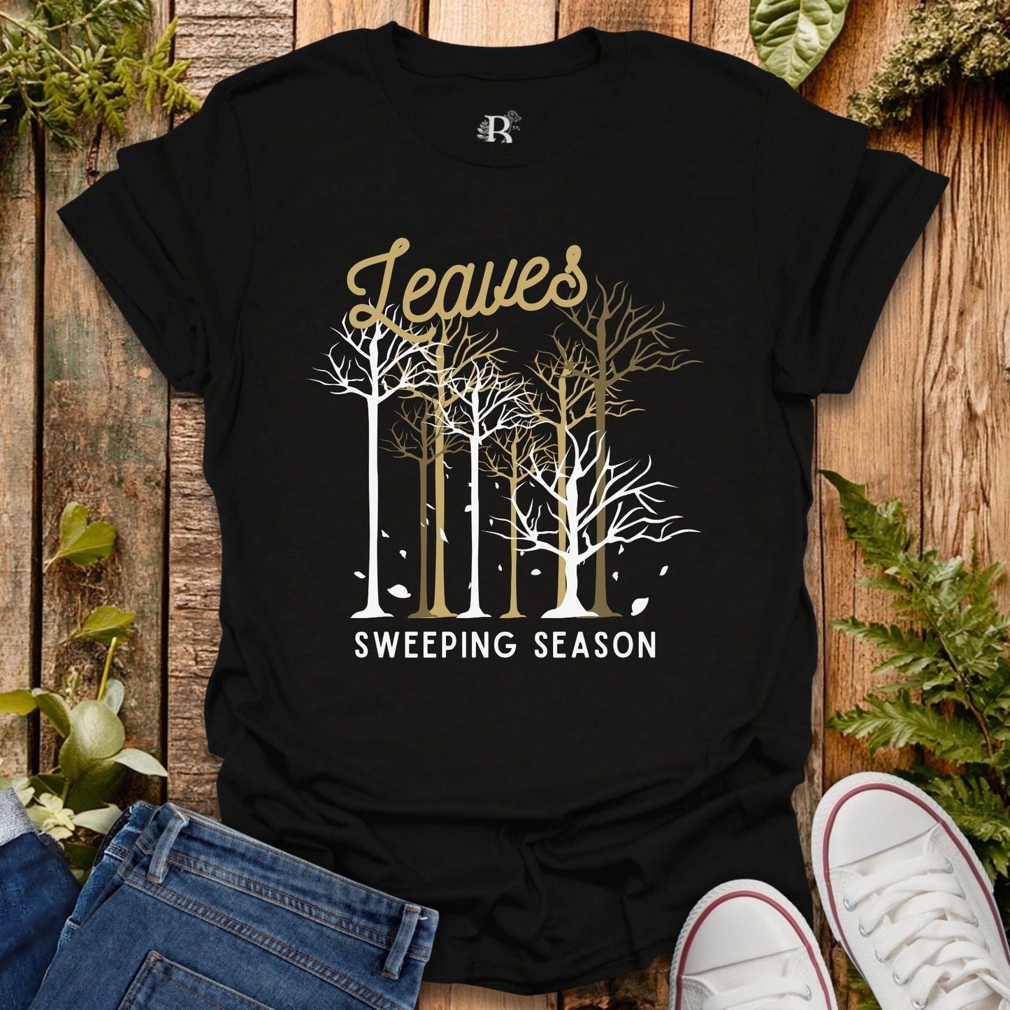 Black t-shirt with 'Leaves Sweeping Season' design on a wooden background with plants.