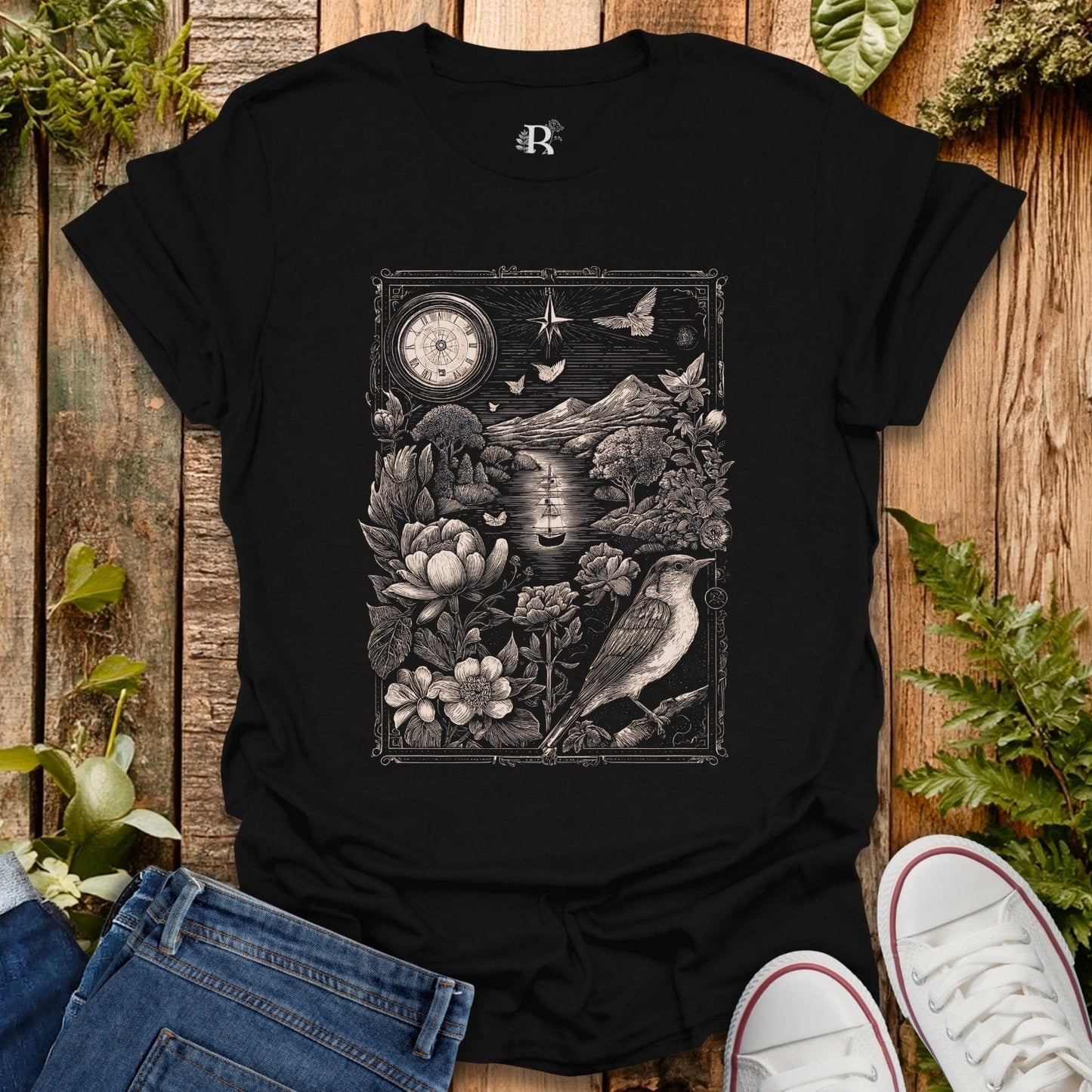 Black t-shirt with intricate design of a bird, flowers, and clock on a wooden surface with plants.